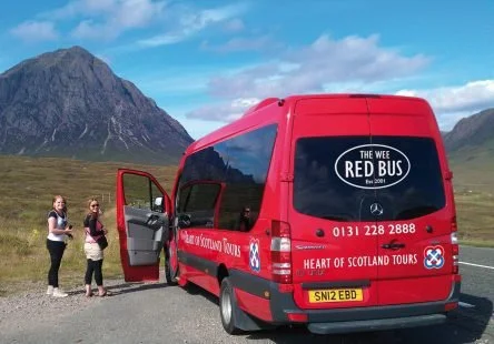 Red touring bus with passengers loading luggage, set against mountains and a blue sky with clouds.