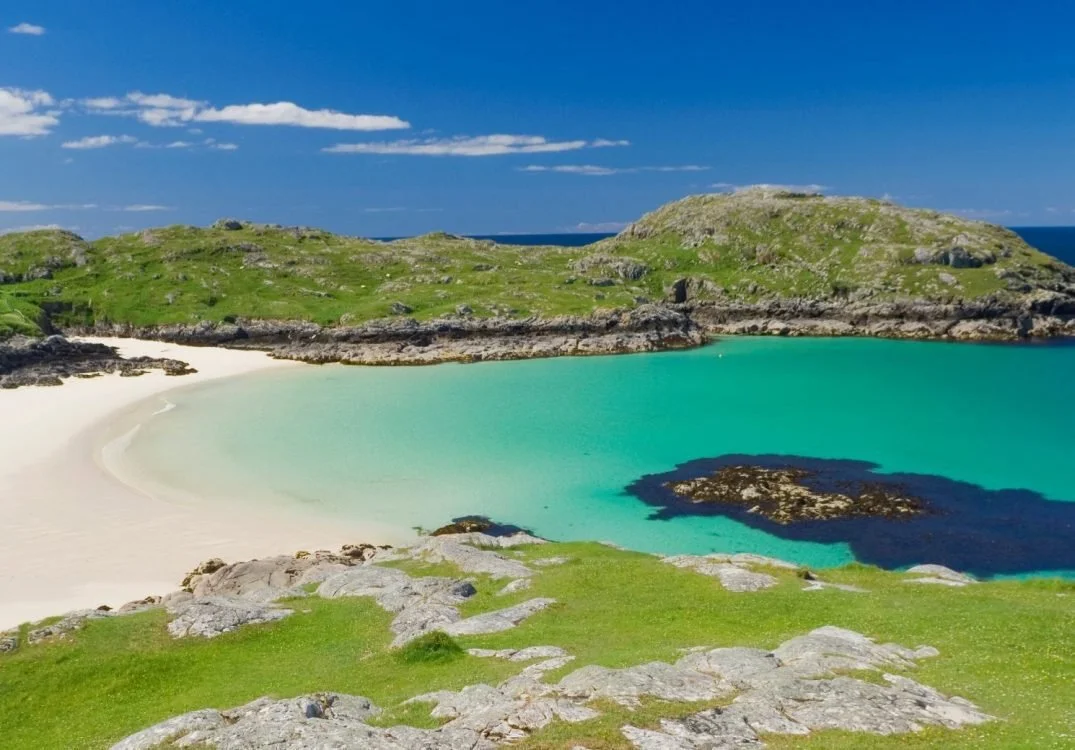 A picturesque beach with white sand, turquoise water, green grassy cliffs, and rocky hills under a blue sky with some clouds.