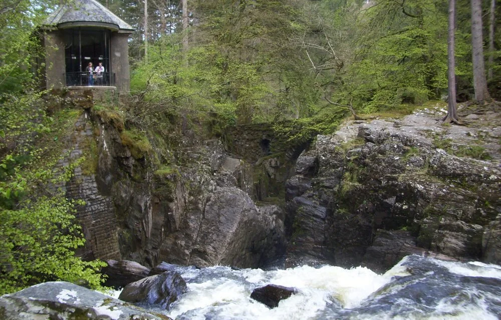 A scenic view of a rocky river flowing through a forest with a stone bridge spanning the gorge. A small observation hut with people inside is situated above the river on the left side.