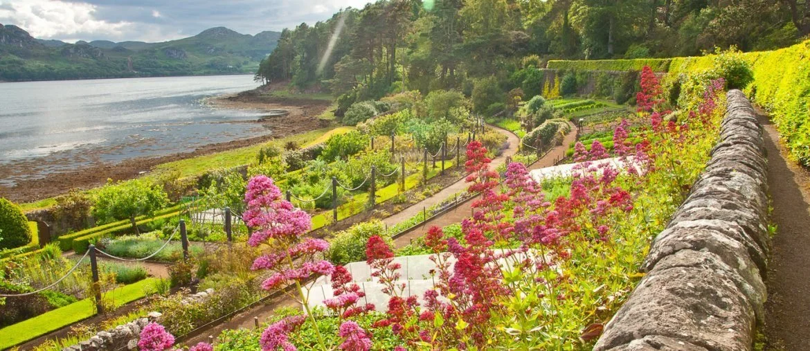 Lush garden with colorful flowers, pathways, and a stone wall overlooking a lake with mountains in the background.