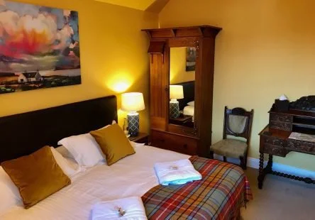 Hotel bedroom with a bed, two pillows, a blanket, a wooden wardrobe, a chair, a dresser, and a colorful painting on the yellow wall.