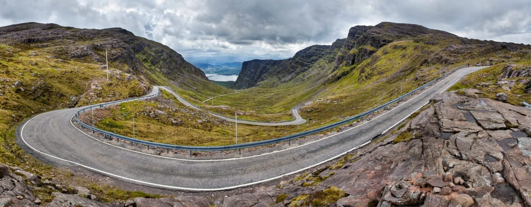 A winding mountain road with guardrails, surrounded by green moss-covered hills and rocky terrain, under a cloudy sky.