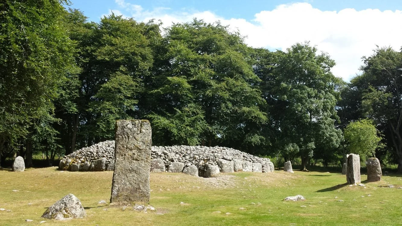 An ancient stone circle with large upright stones on grassy ground, surrounded by trees under a partly cloudy sky.