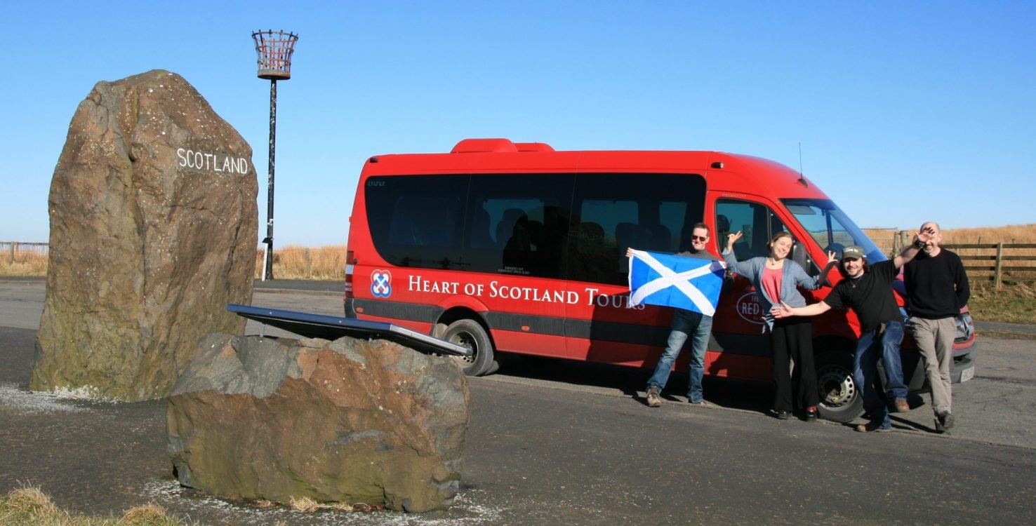 People standing in front of a red tour van with a blue Scottish flag, next to a large rock with 'SCOTLAND' written on it, in a scenic outdoor setting.