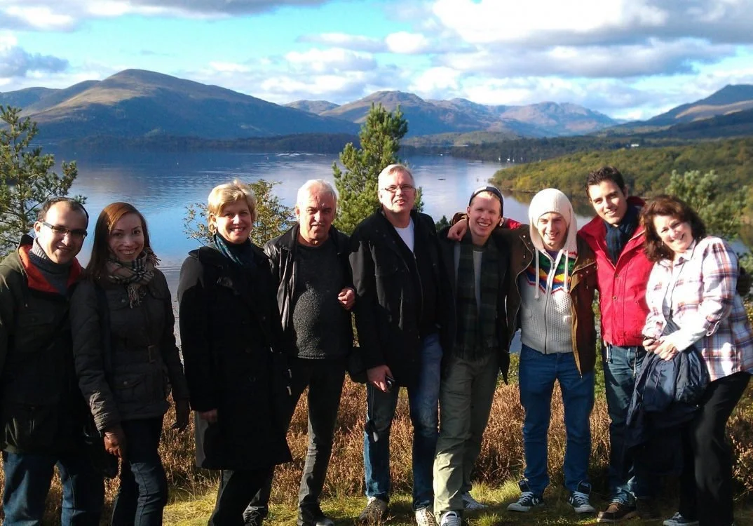 Group of nine people standing outdoors by a lake with mountains in the background, smiling at the camera.