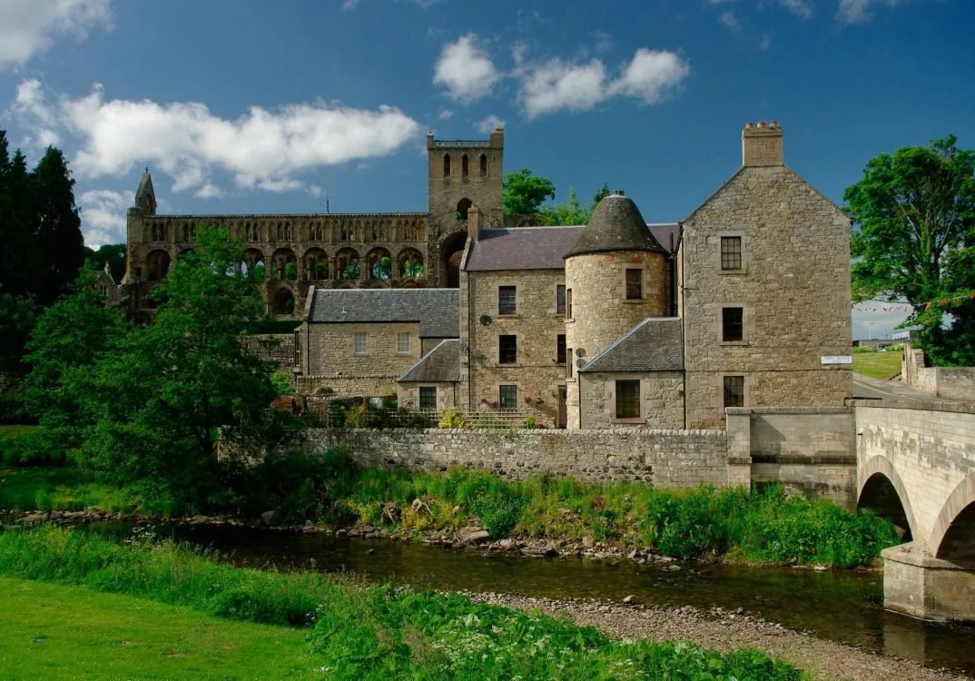 Historical stone buildings and castle ruins near a river with green trees and a bright blue sky.