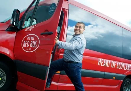 A smiling man in a light blue shirt standing next to a red bus with the logo 'The Wee Red Bus' and the text 'Heart of Scotland' on it.