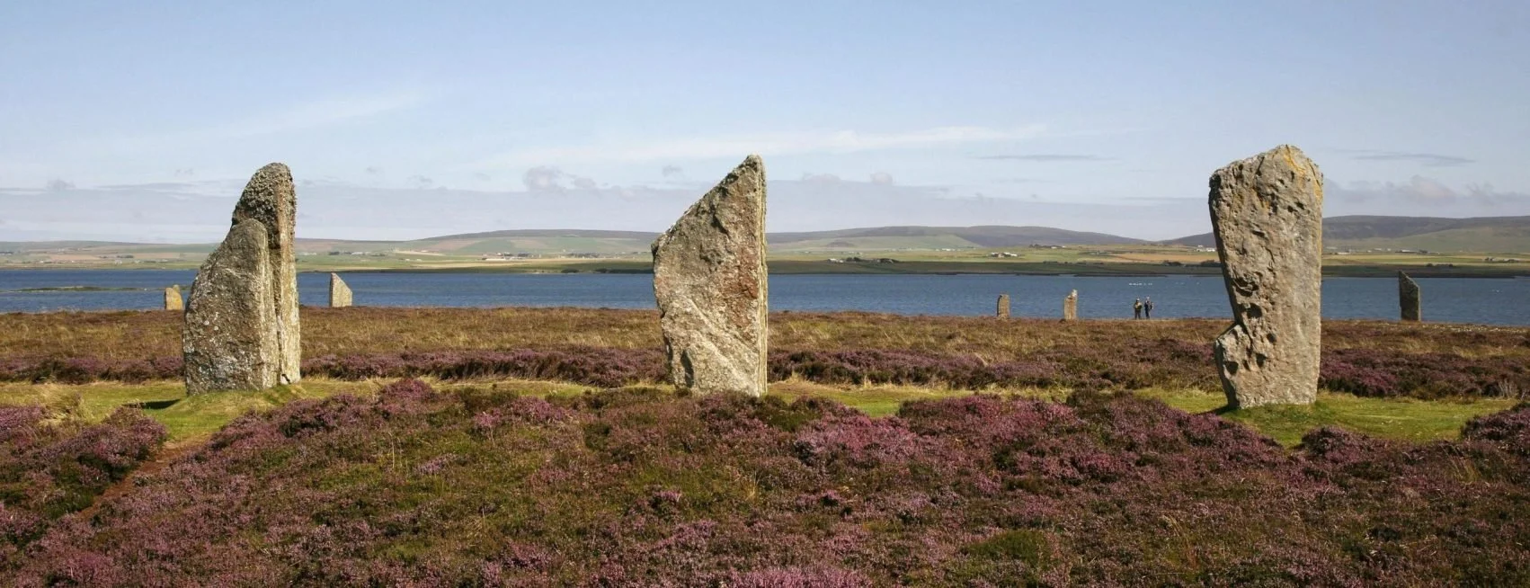 Three standing stones in a grassy and purple-flowered landscape overlooking a body of water with hills in the background.