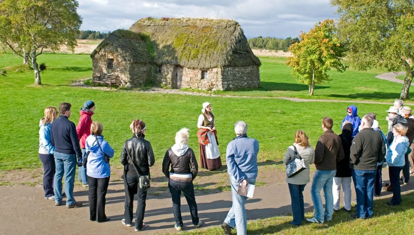 A group of tourists listening to a woman in historical costume giving a tour outside a thatched-roof stone cottage in a grassy area with trees and a cloudy sky.