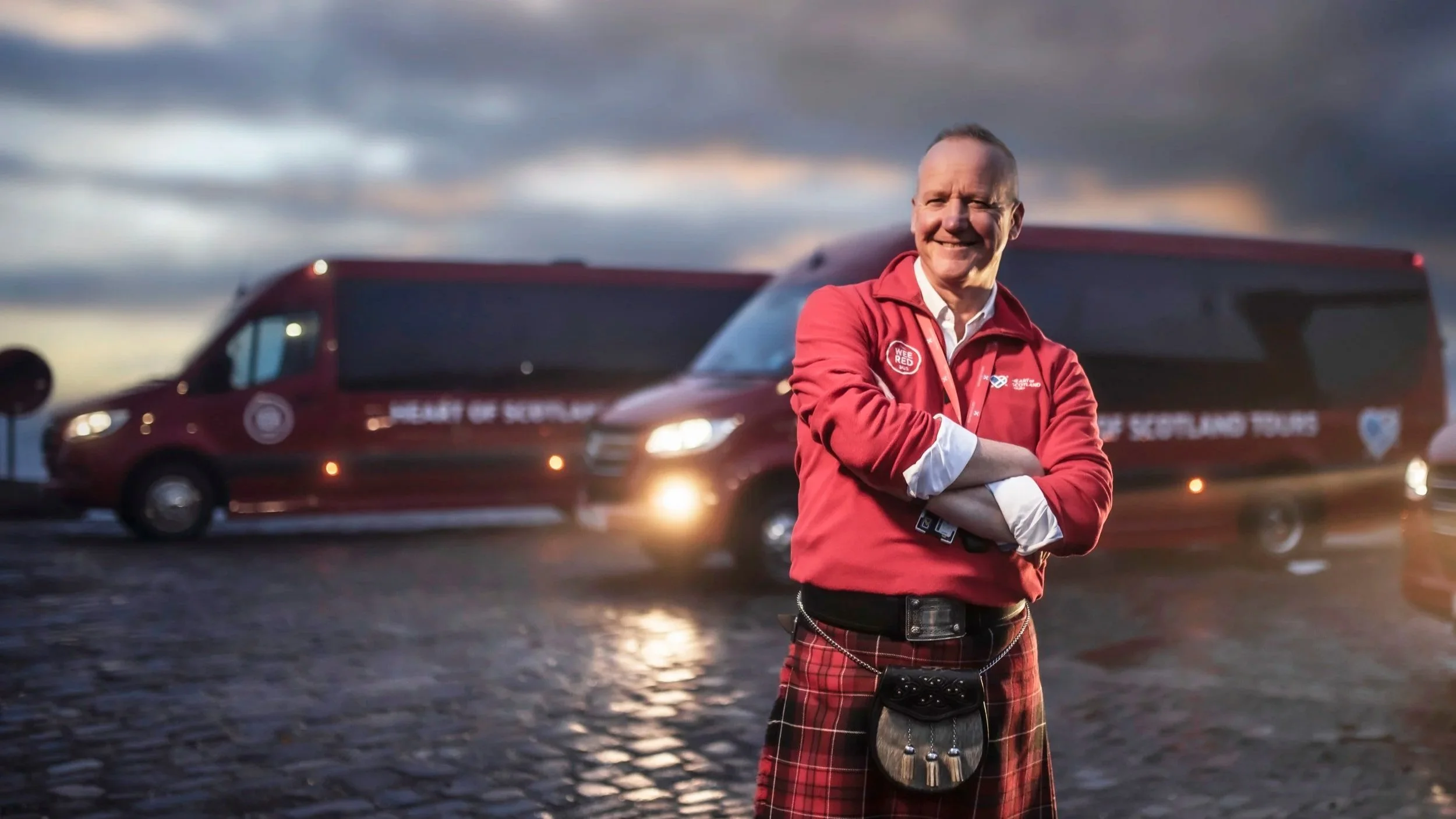 A man wearing a red jacket and plaid kilt standing outdoors with his arms crossed, smiling, with a large red bus in the background on a rainy day.