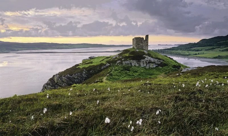 Ancient stone tower on a hill overlooking a wide river with lush green fields and cloudy sky in the background.
