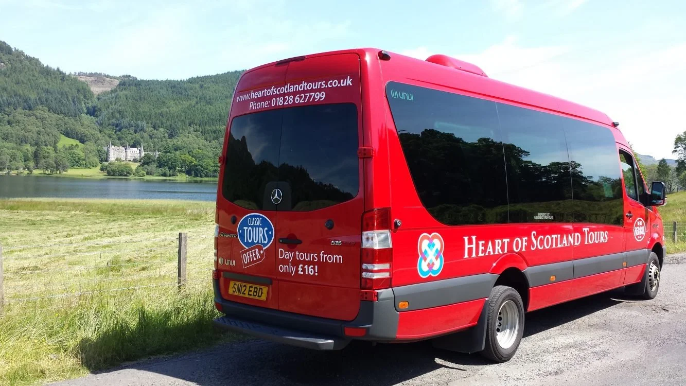 Red tour bus with 'Heart of Scotland Tours' written on the side, parked near a grassy field with a lake and forested hills in the background.