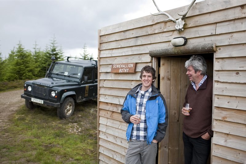 Two men standing outside a small wooden building with a sign that reads 'Schiehallion Bothy,' one holding a drink, with a land rover parked nearby and a deer skull with antlers mounted above the doorway.