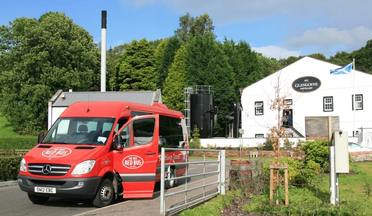 Red tour bus branded 'The Wee Red Bus' parked in front of a white distillery building labeled 'Glengoyne Distillery,' with a blue and white Scottish flag, trees, and a cloudy sky in the background.