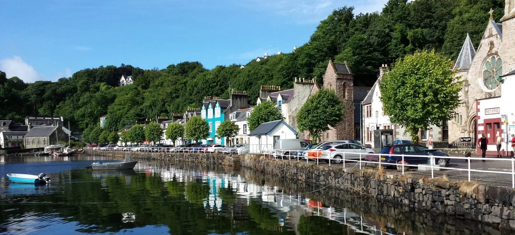 A scenic harbor with small boats anchored in calm water, colorful buildings along the waterfront, parked cars, and a hillside covered in lush green trees in the background.