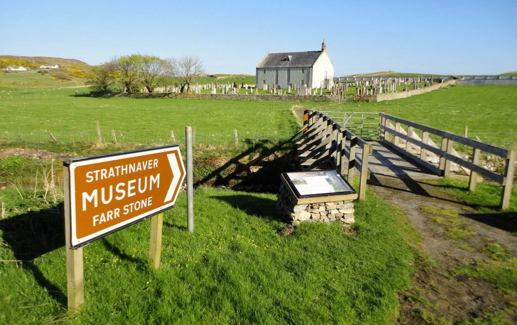 Sign pointing to the Strathnaver Museum and Farr Stone, with a grassy hill, a church, and tombstones in the background under a clear blue sky.