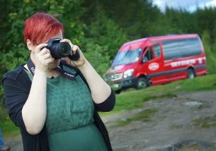 A woman with red hair taking a photo with a camera outdoors, with a red bus in the background near greenery.