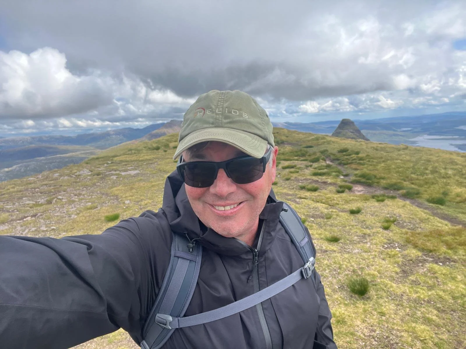 A man in outdoor gear, including a green cap and black sunglasses, taking a selfie on a mountain trail with lush green hills, a prominent mountain peak in the distance, and a cloudy sky overhead.