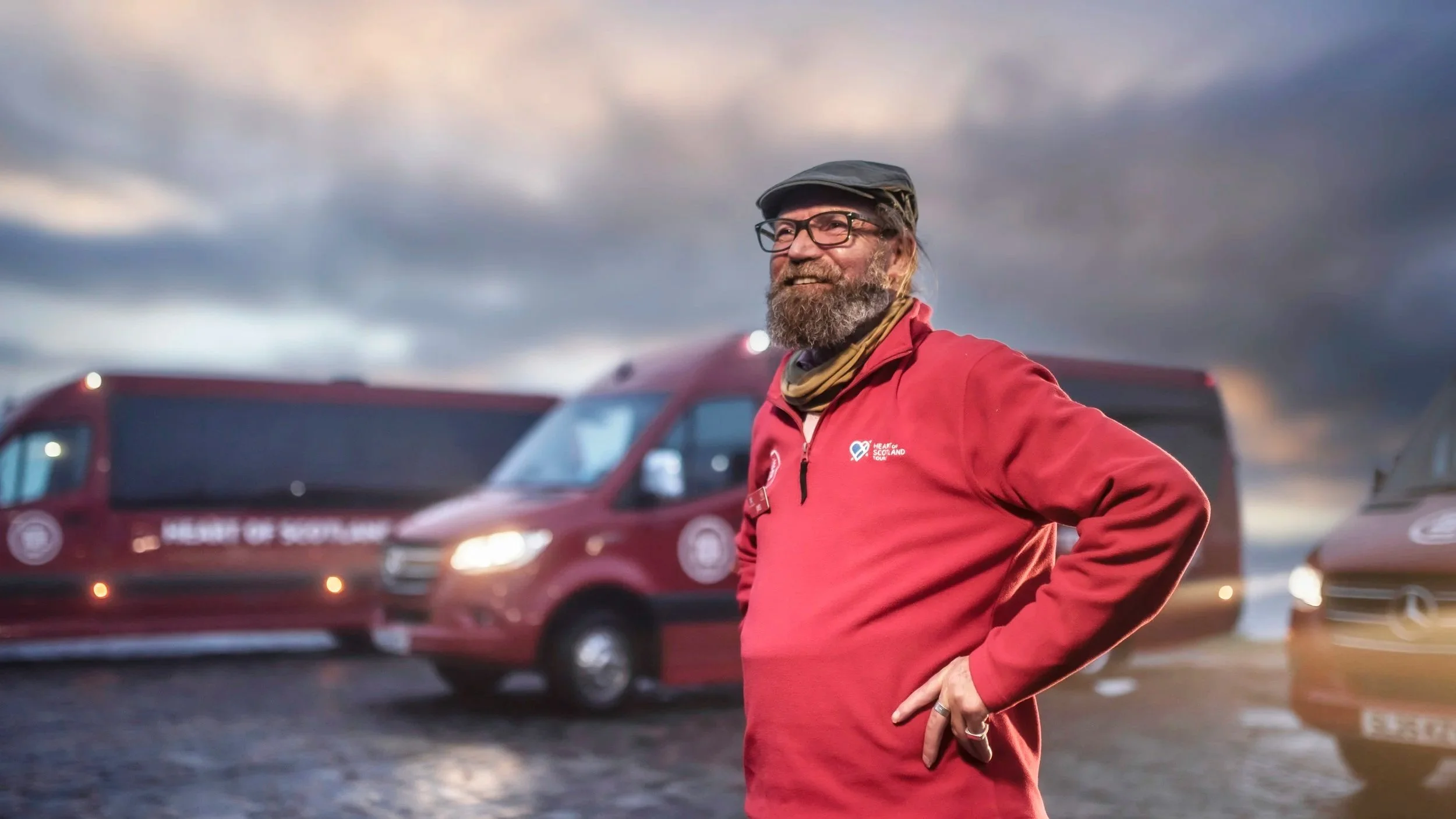 A bearded man wearing glasses, a black cap, and a red jacket with a logo stands outdoors with his hands on his hips, smiling. There are red vans and other vehicles in the background under a cloudy sky, indicating a parking lot or outdoor event setting.