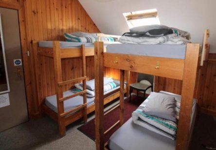 A room with a wooden bunk bed, a single bed, and wood-paneled walls. The bunk bed has pillows and blankets, and there's a small chair next to the single bed.