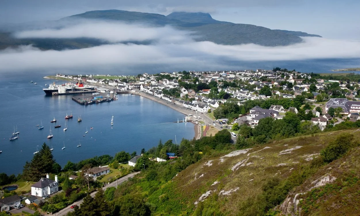 A coastal town with white buildings, a harbor with boats, and a mountain shrouded in clouds in the background.