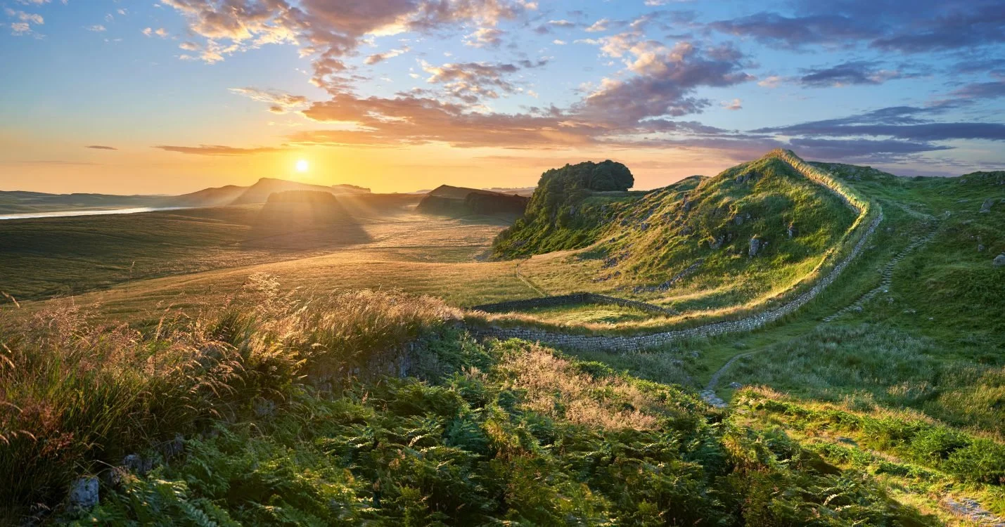 A scenic view of rolling green hills during sunset with a winding stone wall and a dirt path, under a sky with scattered clouds.