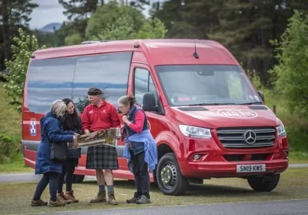 Four people gathered around a map in front of a red Mercedes-Benz van parked outdoors with trees in the background.