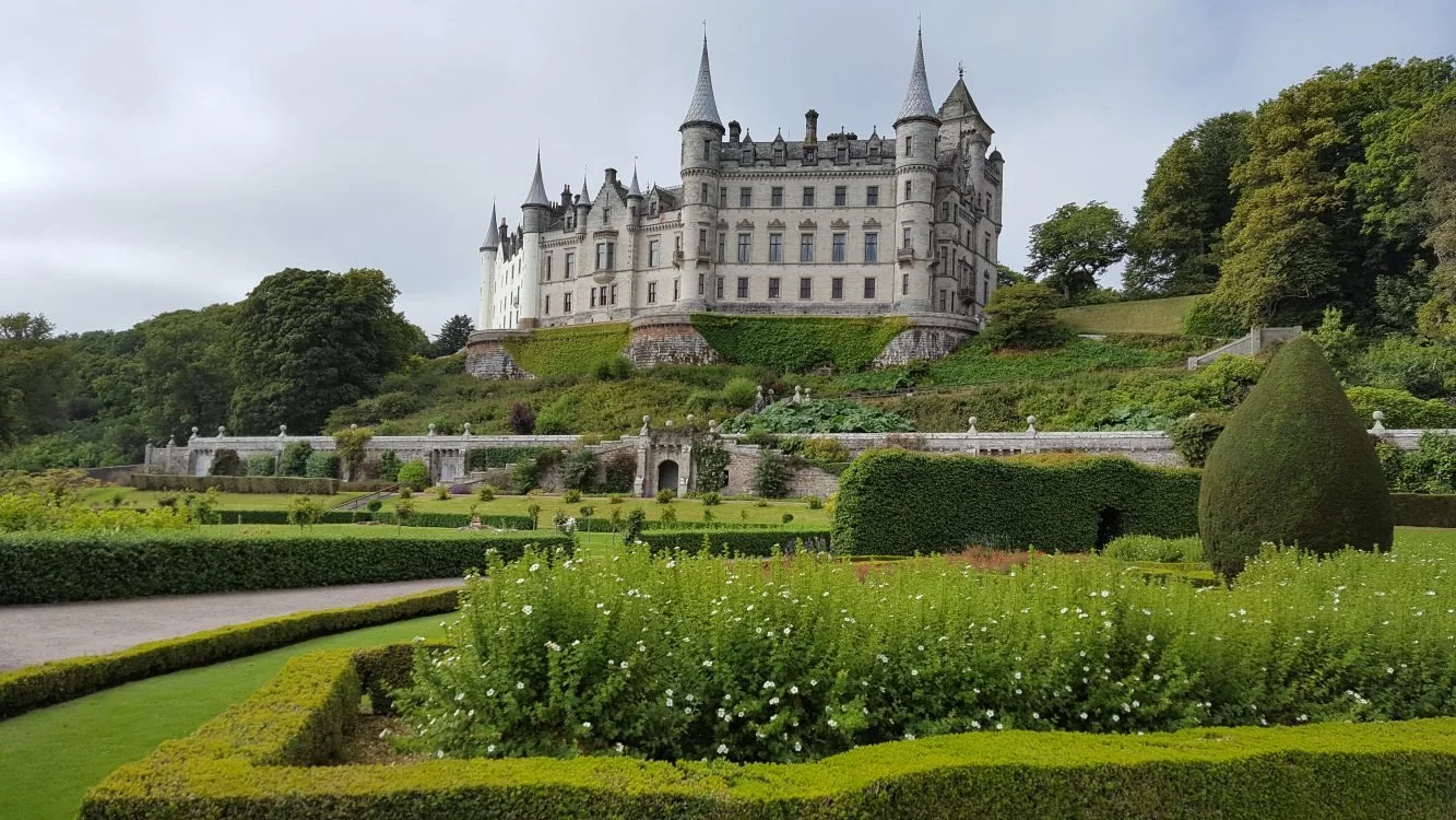 A large, historic castle with multiple turrets sits atop a hill, surrounded by lush gardens and greenery.