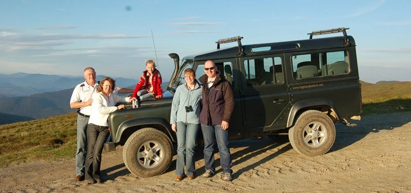 Group of five people posing next to a black off-road vehicle on a hilltop with mountain scenery in the background.