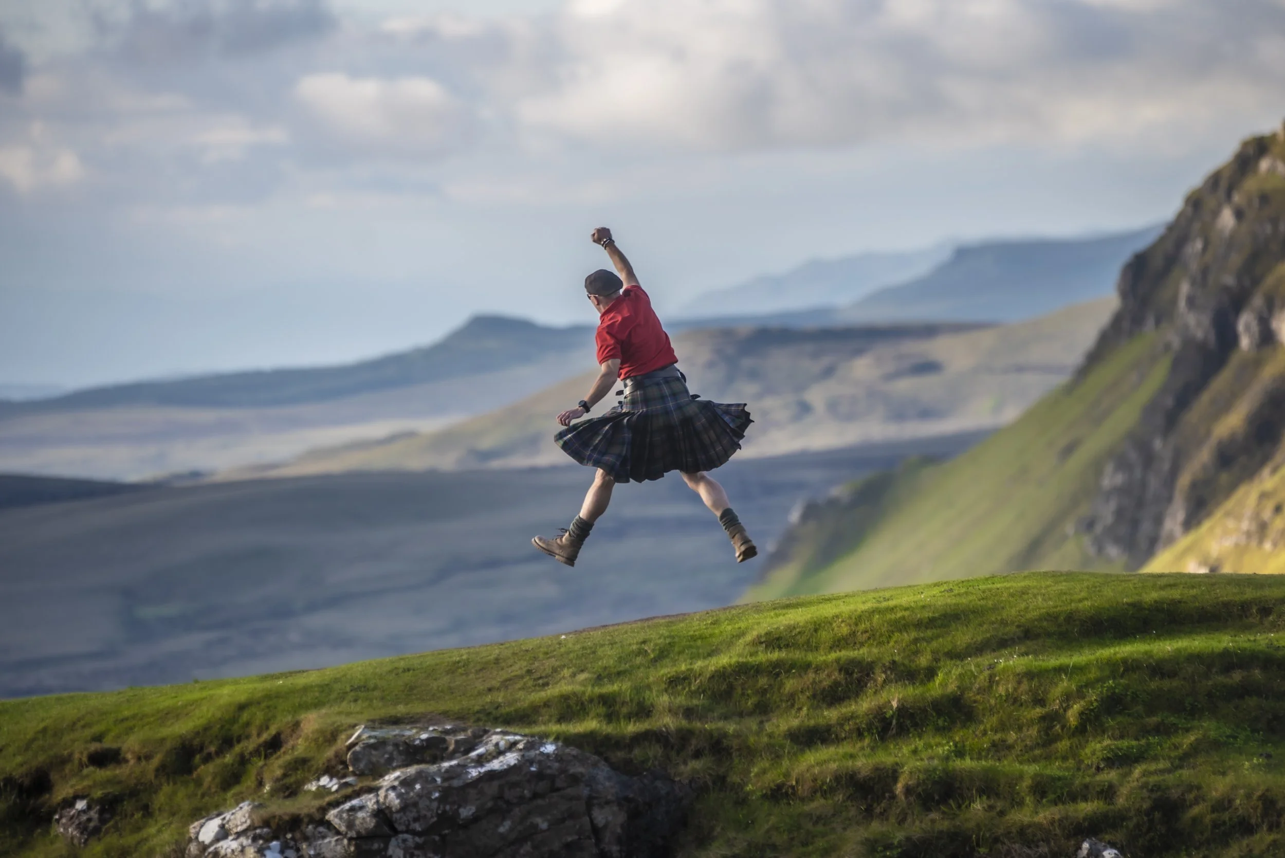 A man dressed in a red shirt, tartan kilt, and hiking boots jumps joyfully across a grassy landscape with mountains in the background.