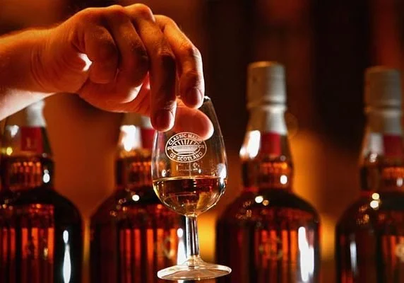 Person holding a small glass of liquor with an emblem, with several larger bottles of liquor in the background.