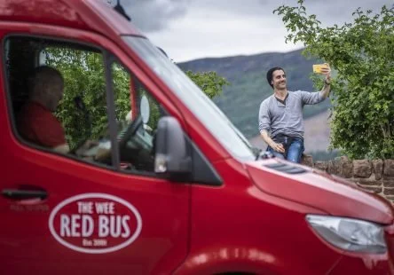 A man is taking a selfie outside near a body of water with trees in the background, while a driver inside a red bus labeled 'The WEEE Red Bus' looks on.