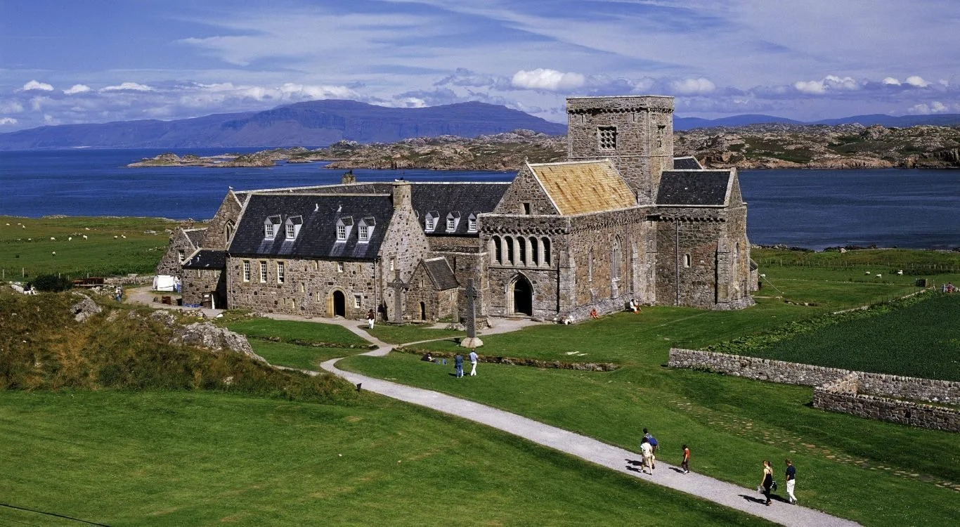 A historic stone church with a tower, surrounded by green fields, near a large body of water with islands and mountains in the background, and people walking along a gravel path.