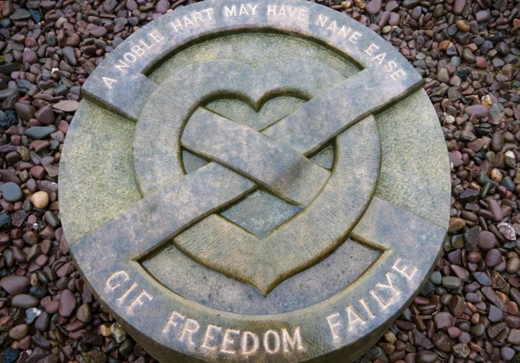 A memorial stone with an engraved heart and ribbon symbol, surrounded by small colored rocks, inscribed with the words 'A Noble Hart May Have Nane, Ease, Cuf, Freedom, Faidye'.