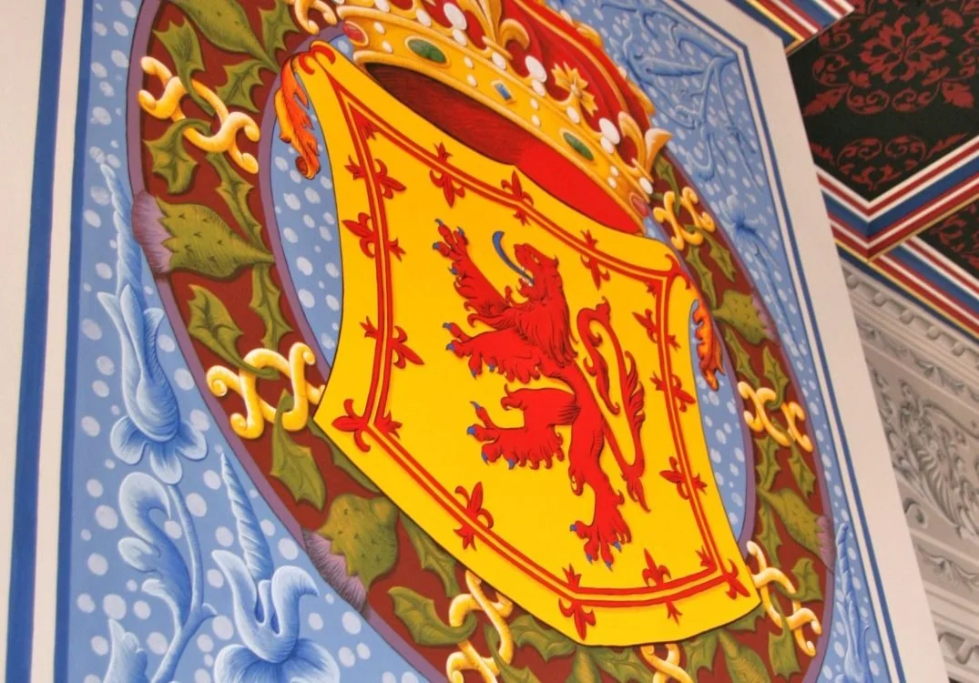 Close-up of a detailed, colorful crest with a yellow shield featuring a red lion, surrounded by green leaves, and topped with a star and crown. The crest is part of a decorative wall with intricate patterns and bright colors.
