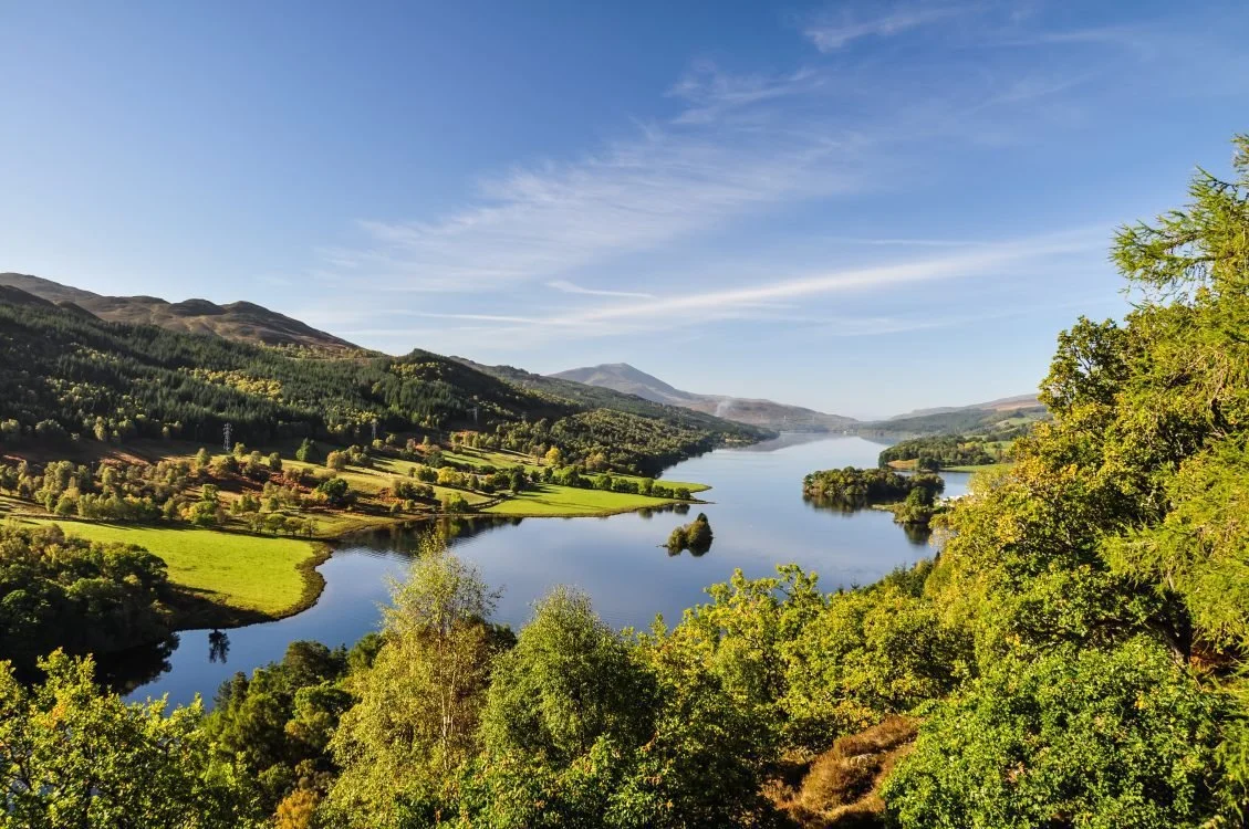Scenic view of a large lake surrounded by green trees, rolling hills, and mountains under a partly cloudy sky