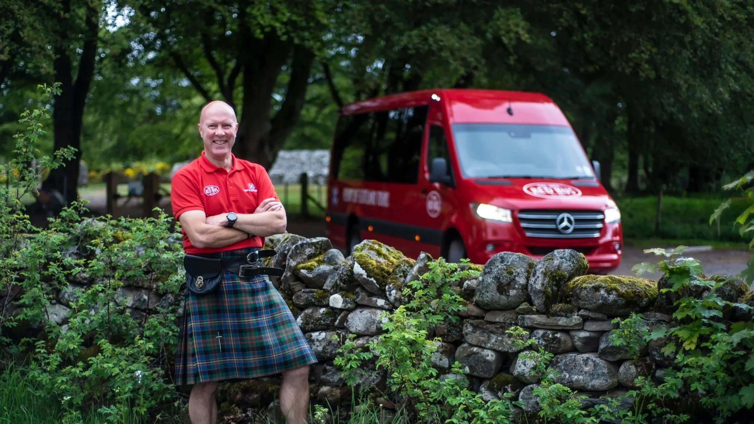 A man in a red shirt and tartan kilt smiling with arms crossed next to a stone wall, with a red Mercedes-Benz emergency response vehicle in the background in a park with trees.