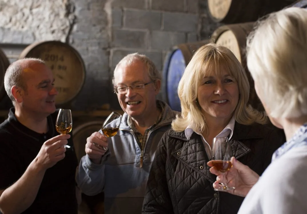 Four adults holding glasses of whiskey in a cellar with barrels in the background, engaged in conversation, smiling and toasting.