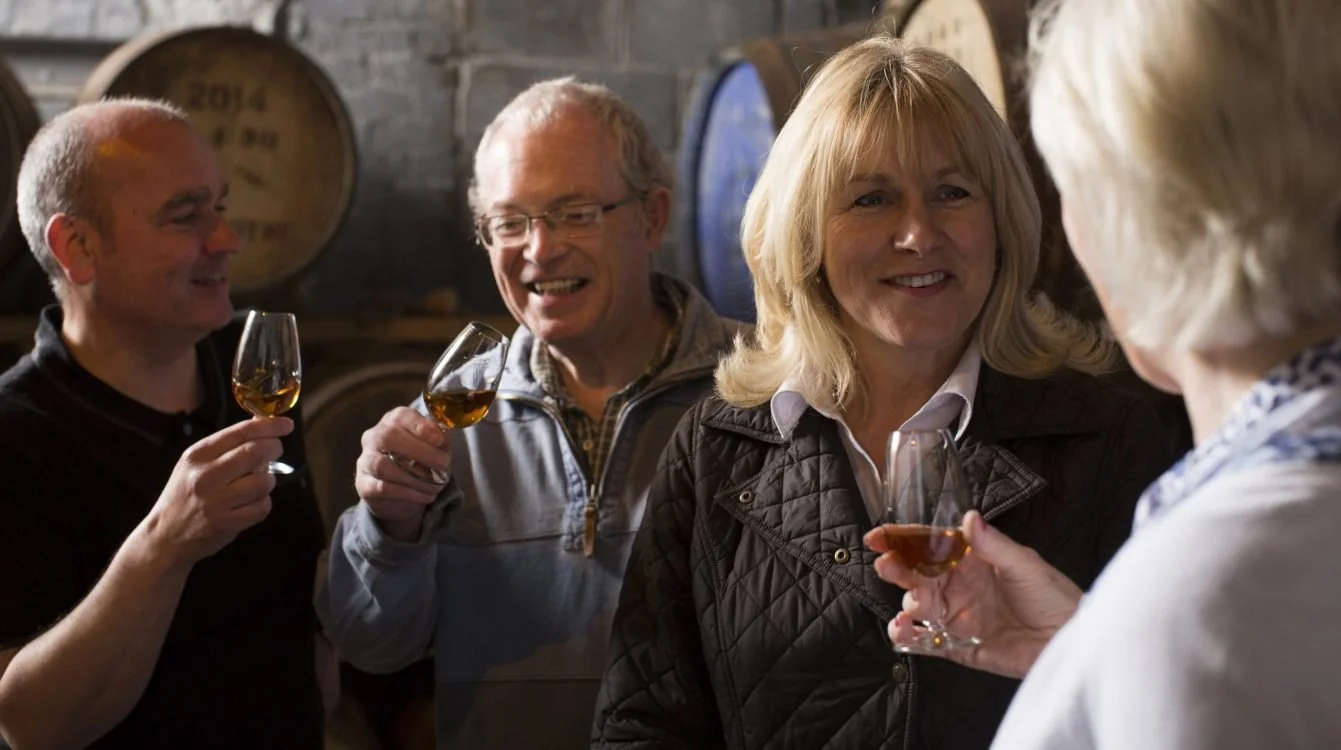 Four adults sampling whiskey in a distillery or cellar, holding glasses of amber-colored whiskey and engaging in conversation.