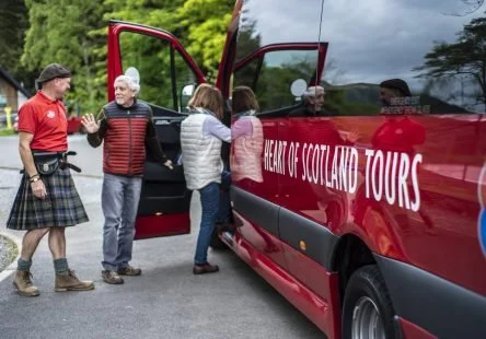 Group of people at a tour bus stopped outdoors with open doors, engaging in conversation. One person is dressed in a kilt. The bus has text that reads 'Heart of Scotland Tours'.