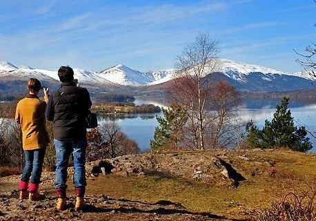 Two people standing on a grassy area near a lake with snow-capped mountains in the background, trees, and clear blue sky.