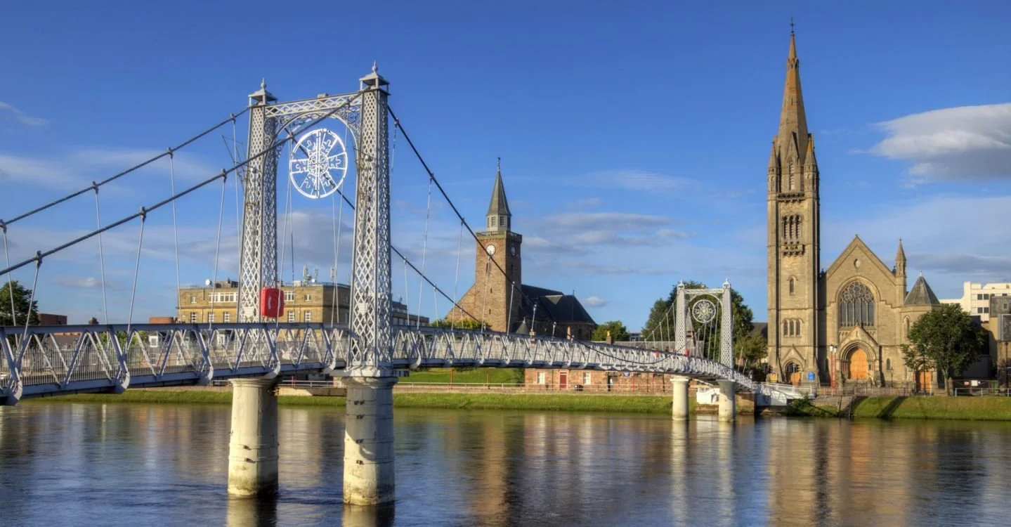 A suspension bridge over a river with a church with a tall steeple in the background, under a partly cloudy blue sky.