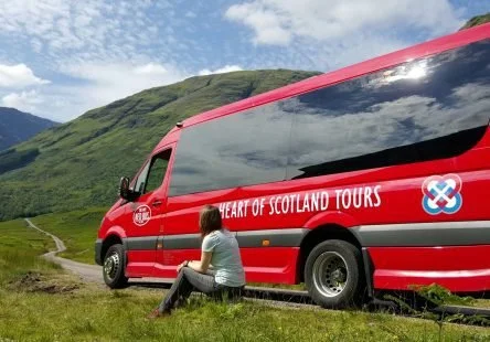 A woman sitting on the grass beside a red tour bus that reads 'Heart of Scotland Tours' in a scenic rural landscape with green hills and a cloudy sky.