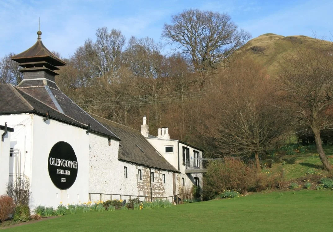 A white building with a black round sign reading 'GLENGOYNE DISTILLERY 1833,' situated on a grassy area with blooming flowers. There are trees and a hill with sparse vegetation in the background under a blue sky.