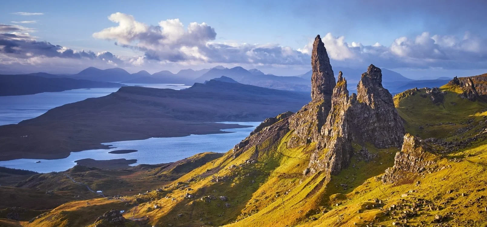 A scenic landscape of rugged jagged rock formations on a grassy hillside with lakes and mountains in the distance under a partly cloudy sky.