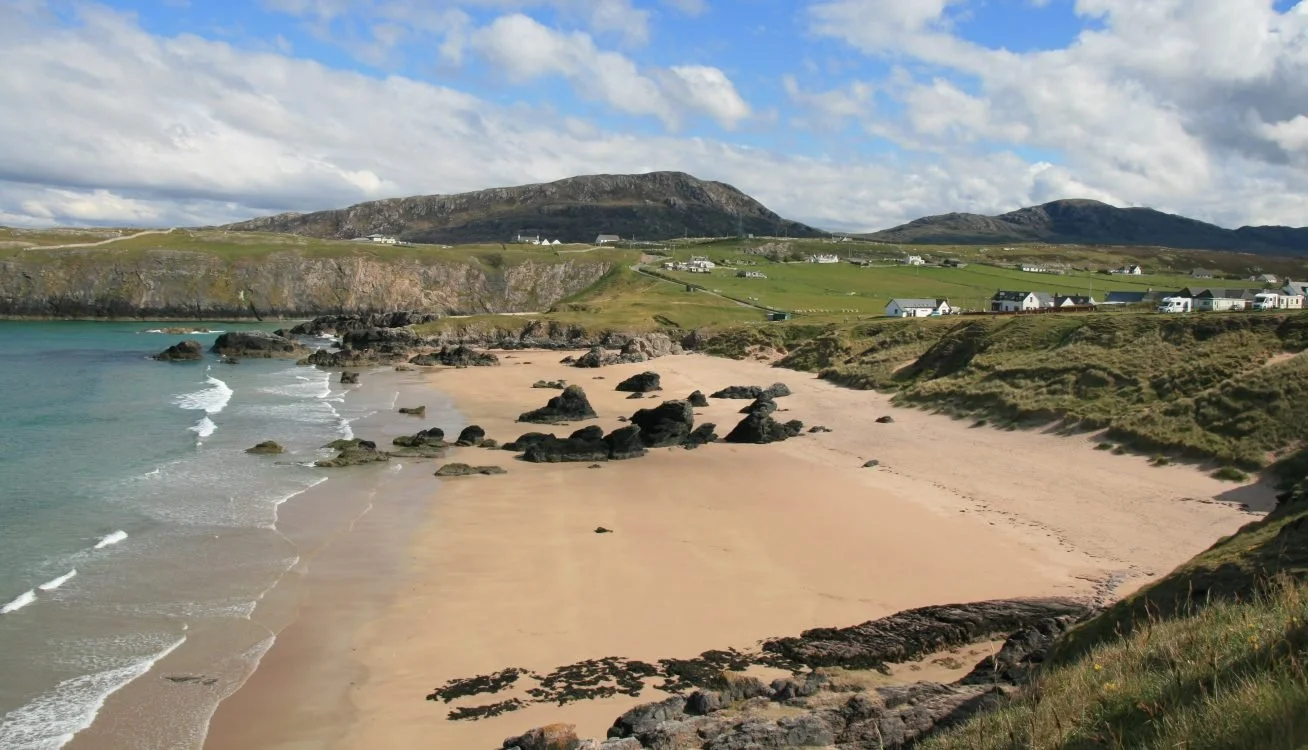 A beach with sand, rocks, and waves, surrounded by hillside grass and green fields, with houses and mountains in the background under a partly cloudy sky.