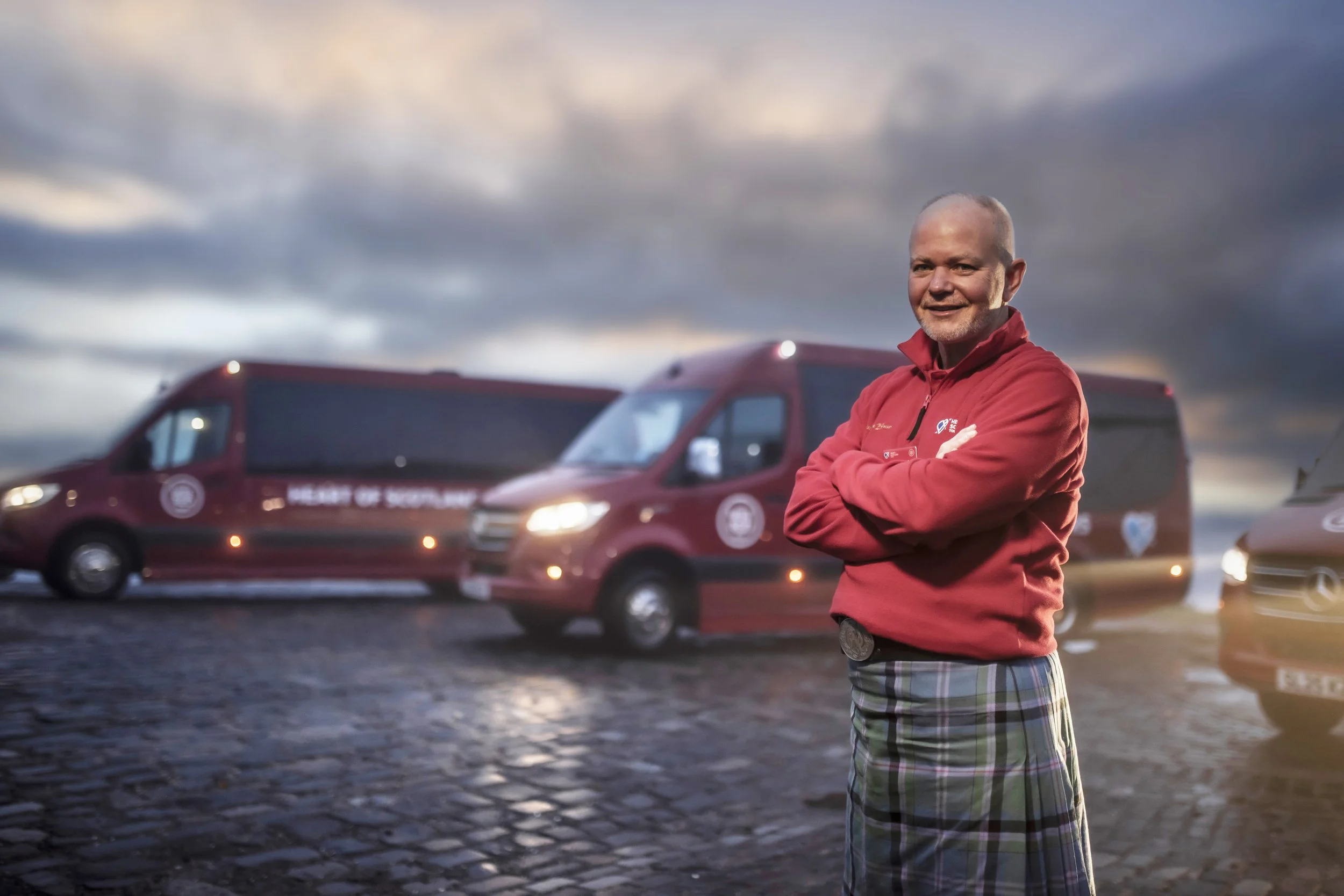 A smiling man with a bald head and beard wearing a red jacket and plaid shorts standing outdoors with cross arms, muddy ground, and red vehicles in the background during cloudy weather at sunset.
