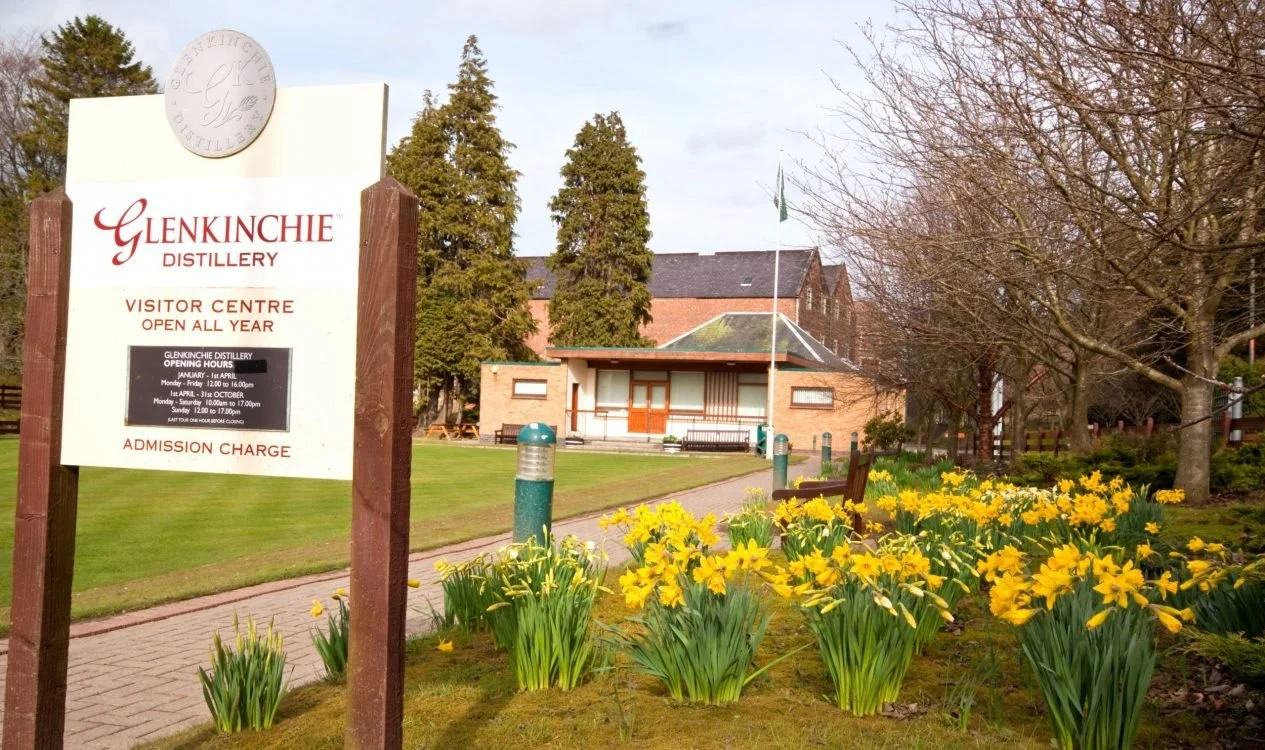 Sign for Glenkinchie Distillery Visitor Centre surrounded by yellow flowers, trees, and a building in the background on a clear day.
