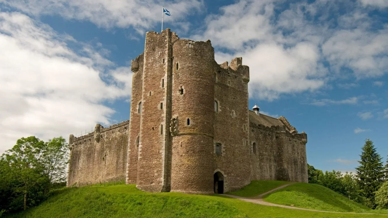 A large stone castle with a Scottish flag on top, surrounded by green grass and trees, against a partly cloudy sky.