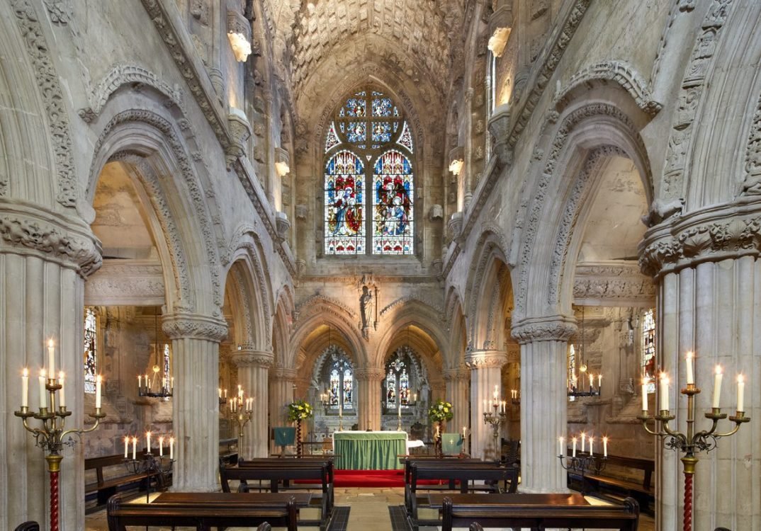 Interior of a historic church with stone walls, ornate arches, stained glass windows, and lit candelabras along the sides, with an altar decorated with flowers at the front.